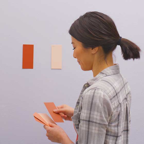 Woman holds henna- and cream-hued swatches with two swatches on the wall as she ponders her choices.