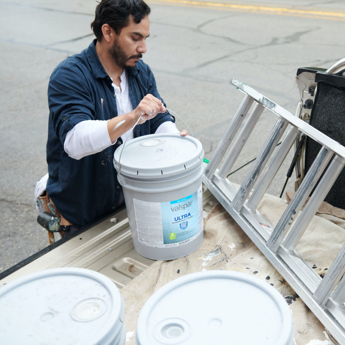 Pro painter loads 5-gallon Valspar® Ultra® onto drop cloth onto bed of pickup truck next to a  ladder.