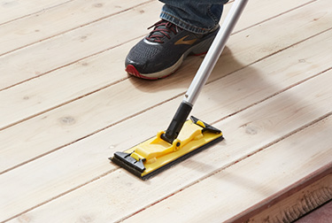 Person in jeans and sneakers sanding wood surface with sander on pole.