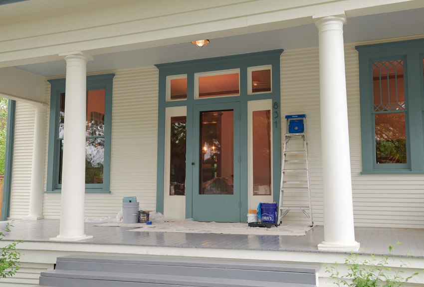 Front entrance to creamy white home. Ladder, drop cloth and paint supplies next to gray-trimmed doors and window.