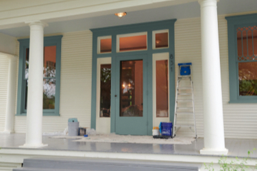 Front entrance to creamy white home. Ladder, drop cloth and paint supplies next to gray-trimmed doors and window.