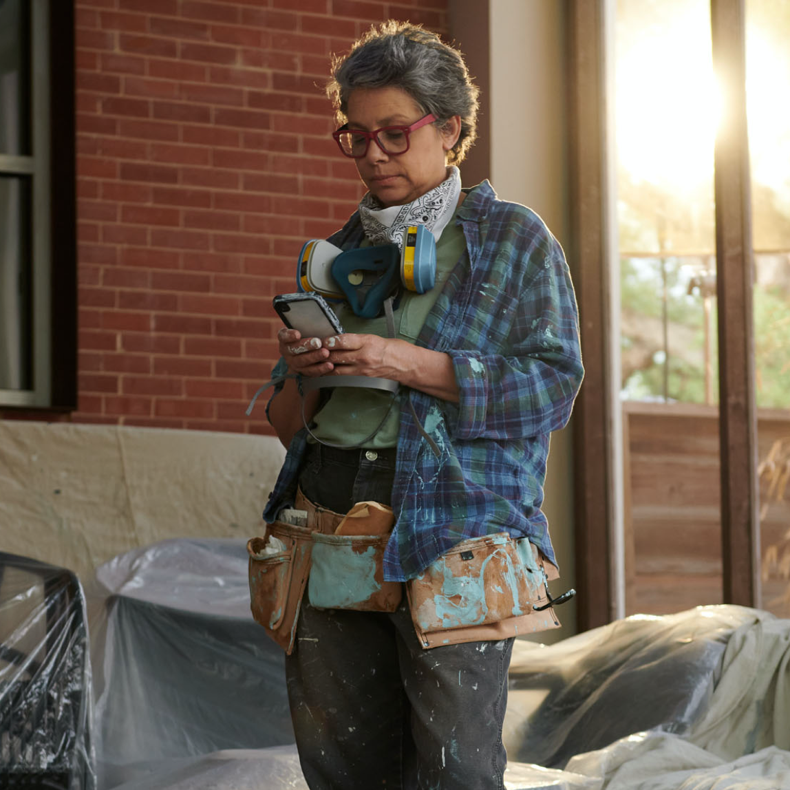 Person wearing paint-stained clothing, using cell phone, with plastic furniture covering in background.