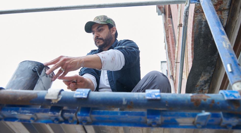 Person on a scaffold in front of a brick building dipping a brush into a can.