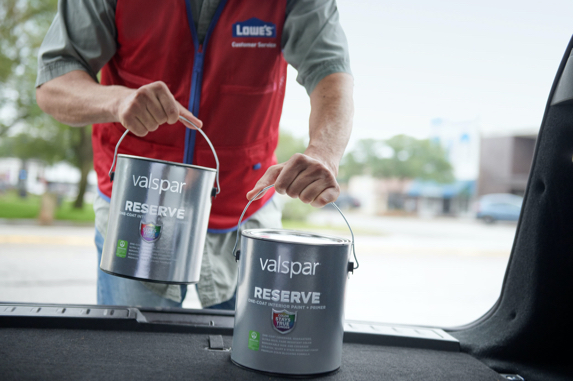 Two cans of paint being loaded into the back of a car by a person in a red vest. 