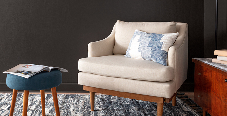 Upholstered chair and four-legged stool against a moody Fired Earth wall. Pillow on chair and open magazine on stool.