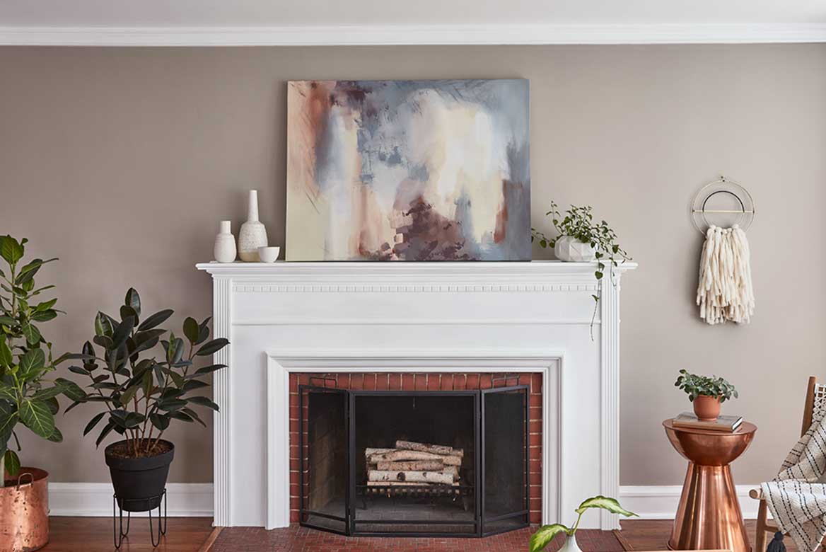 White fireplace against warm wall, copper table and planter and modern art canvas resting on mantle.