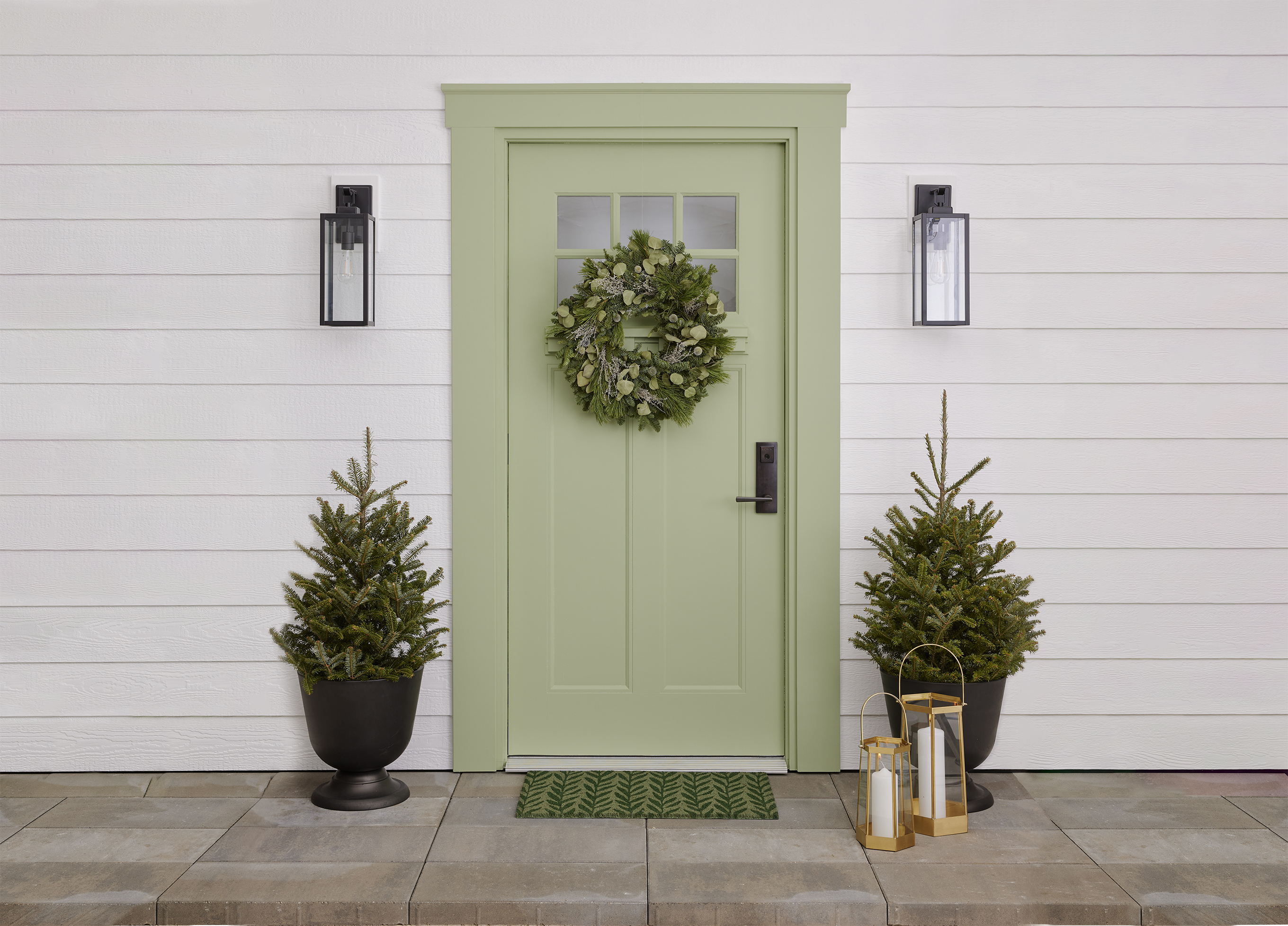 Pale-green door with wreath and doormat. Two potted evergreens and gold lanterns frame the door.