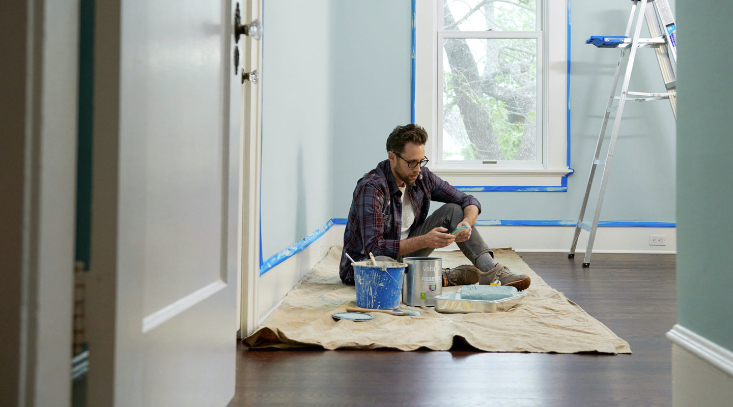 View through doorway of man with paint and tray on drop cloth looking at his phone. The wall is taped off and light blue.