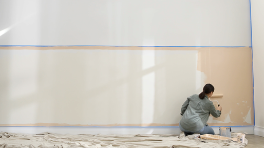 Person sitting down and using a roller on a large taped-off wall with drop cloth on floor.