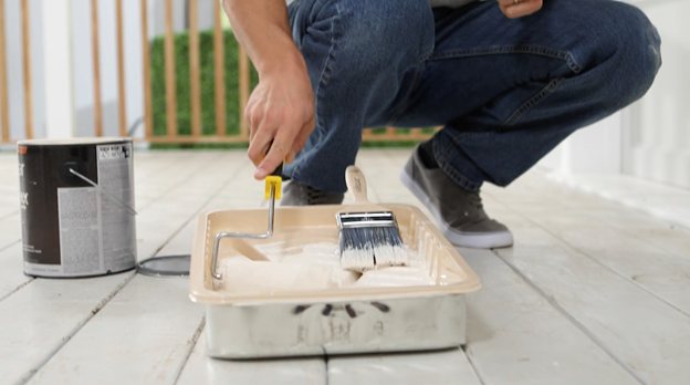 Man is crouched down next to paint tray applying paint to his roller. Brush is in paint tray and can in background.